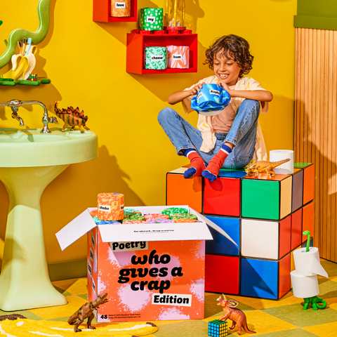 Child sitting on a large Rubik’s cube unwrapping Who Gives A Crap toilet paper